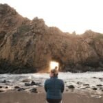 man in blue and black jacket sitting on rock formation in front of sea during daytime